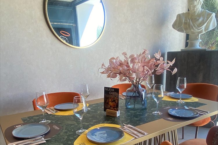 Mesa preparada con vajilla azul, copas y un jarrón de flores bajo un espejo redondo