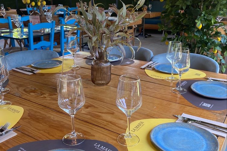 Mesa larga de madera con manteles amarillos, platos azules y sillas azules al fondo